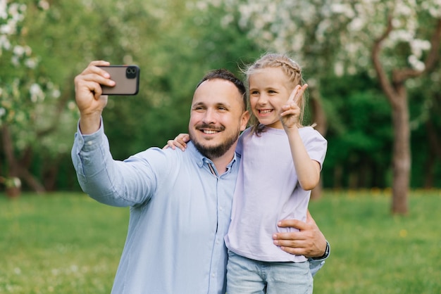 Caucasian dad and daughter smile and take a selfie on their phone while walking in a summer park