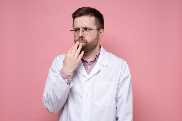 Caucasian doctor in a white coat pensively and anxiously looks to the side holding hand to face