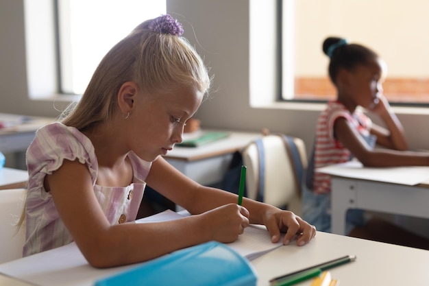 Caucasian elementary schoolgirl writing while studying at desk in classroom