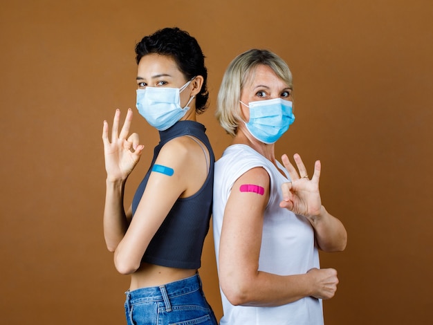 Caucasian female patients wears face mask standing look at camera lean on each other back showing okay or OK hand sign at colorful plaster together after vaccination in front brown background.