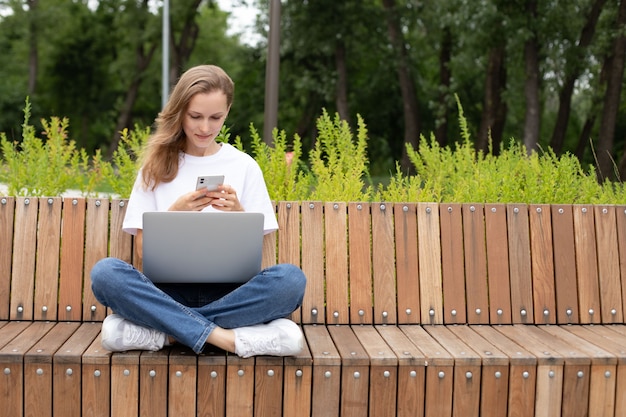 Caucasian female in trendy wear spending time in the park on a wooden bench with phone in hand and laptop. skilled freelancer enjoying remote job typing publication for web blog on smartphone