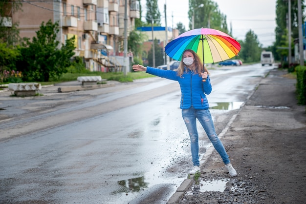 Caucasian girl in a protective mask hails taxi on an empty street, stands with an umbrella in spring rain and waits for car. Safety and social distance during coronavirus pandemic.