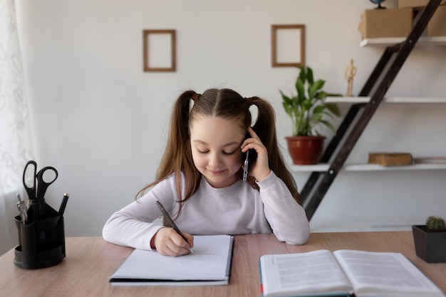 Caucasian girl schoolgirl sits at home at a desk Talking on the phone and making notes in a notebook