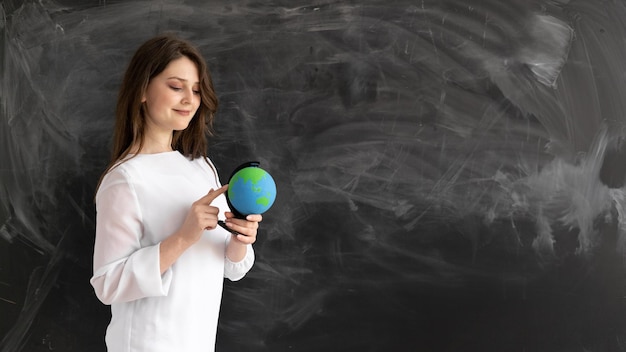 Caucasian girl student stands against the background of a clean chalk board He holds a globe in his hands and points his finger at it Back to school concept geography lesson Baner