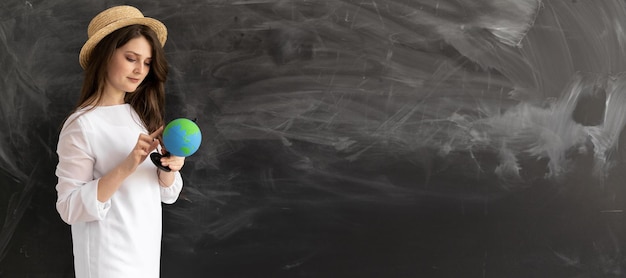 Caucasian girl in a summer hat with a globe standing near an empty chalk board Summer vacation planning concept Copy space