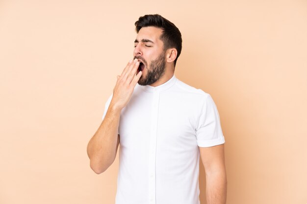 Caucasian handsome man on beige wall yawning and covering wide open mouth with hand