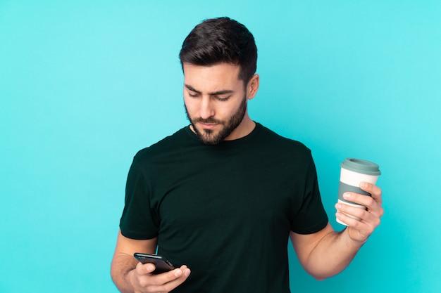 Caucasian handsome man on blue wall holding coffee to take away and a mobile