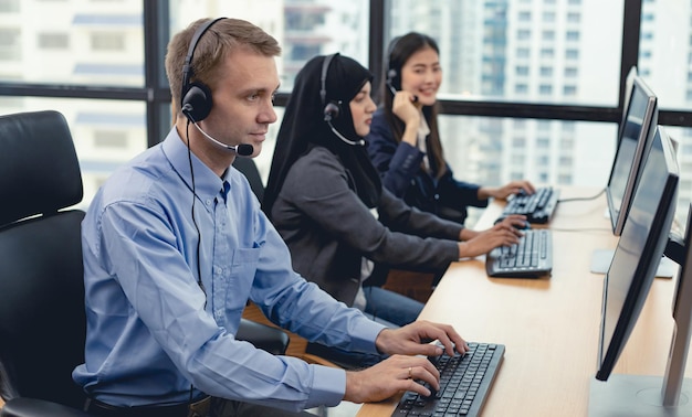 Caucasian handsome man and group of diverse telemarketing customer service staff team in call center. Call center worker accompanied by team. Smiling of customer support operator at work.