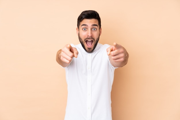 Caucasian handsome man isolated on beige wall surprised and pointing front