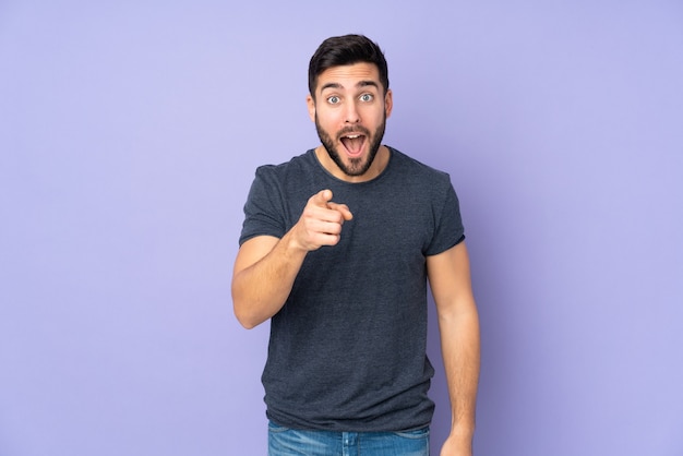 Caucasian handsome man surprised and pointing front over isolated purple wall