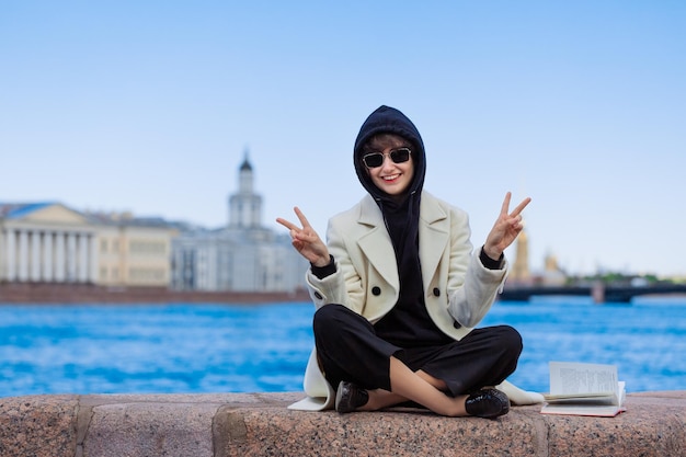 Caucasian happy girl sits on granite curb city with book In a white coat