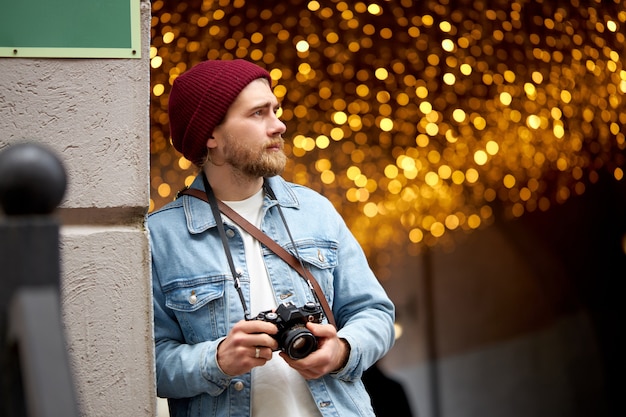 Caucasian hipster guy in denim jacket and red hat walk in city street taking photo, using film camera, Garlands lights in the background, copy space. Photography, people lifestyle concept.