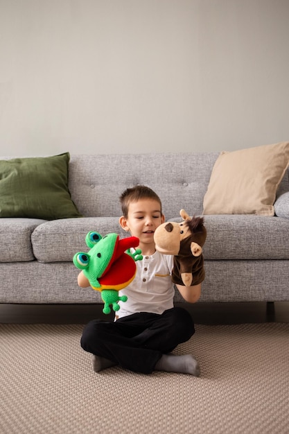 A Caucasian little boy is sitting on the carpet near the sofa and playing a puppet theater with a monkey and a frog