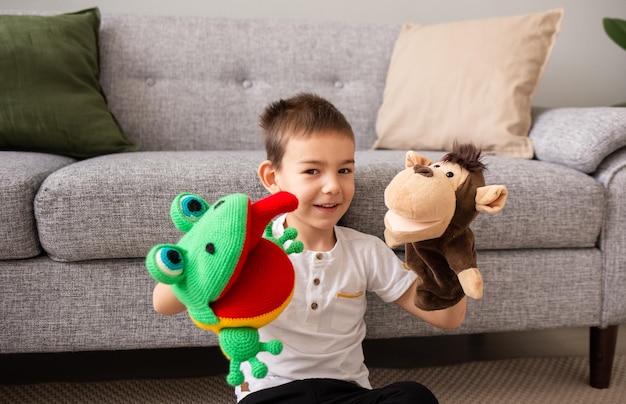 A Caucasian little boy is sitting on the carpet near the sofa in the room and playing a puppet theater with a toy monkey and a frog