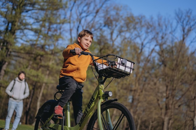 Caucasian little boy riding a bicycle with basket Father walking in distance