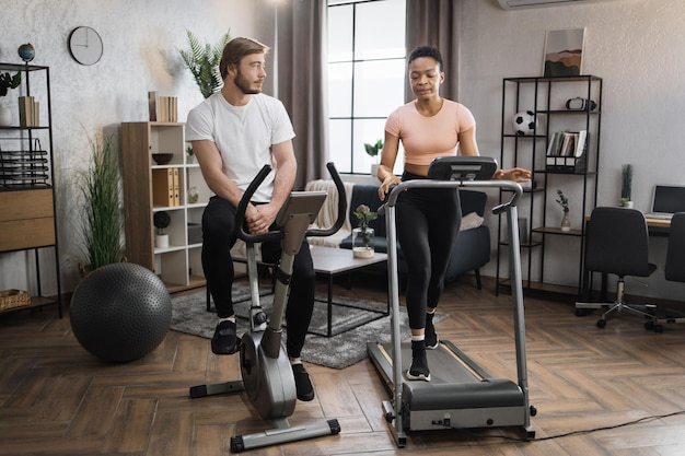 Caucasian male and african female wearing sportswear using exercise bike and treadmill