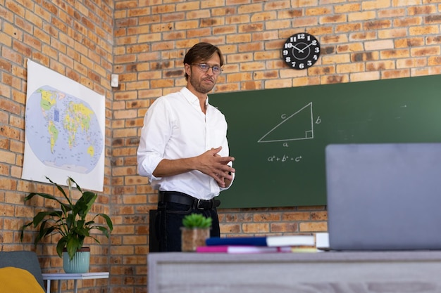 Caucasian male maths teacher standing by blackboard giving online lesson using laptop