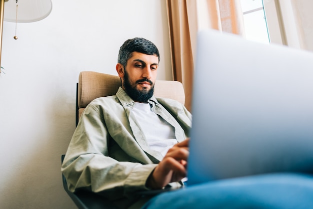 Caucasian man freelancer working at home, sitting on armchair and using laptop computer.