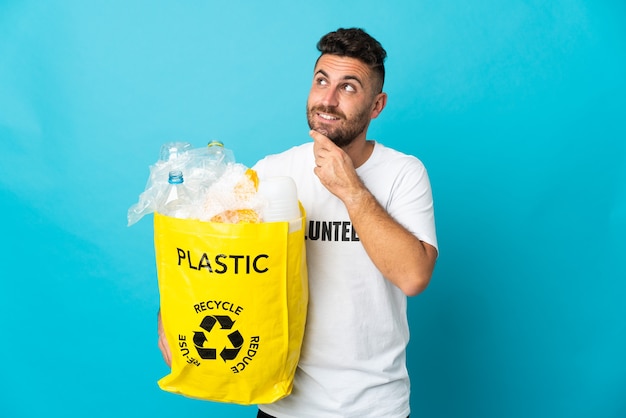 Caucasian man holding a bag full of plastic bottles to recycle isolated on blue background looking up while smiling