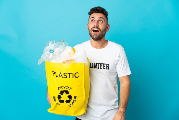 Caucasian man holding a bag full of plastic bottles to recycle isolated on blue background looking up and with surprised expression