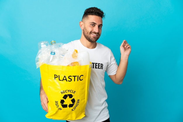 Caucasian man holding a bag full of plastic bottles to recycle isolated on blue background pointing back