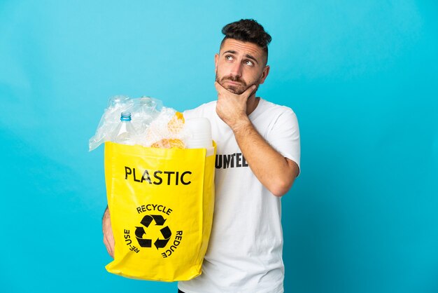 Caucasian man holding a bag full of plastic bottles to recycle isolated on blue having doubts