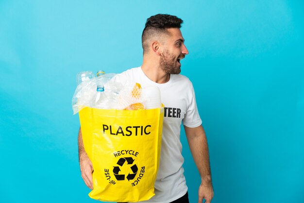 Caucasian man holding a bag full of plastic bottles to recycle isolated on blue wall laughing in lateral position