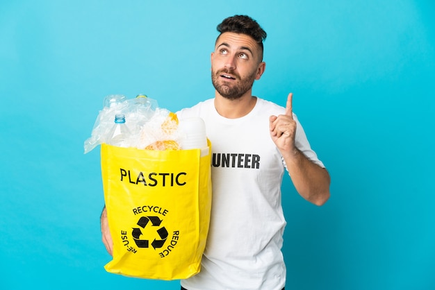 Caucasian man holding a bag full of plastic bottles to recycle isolated on blue wall thinking an idea pointing the finger up