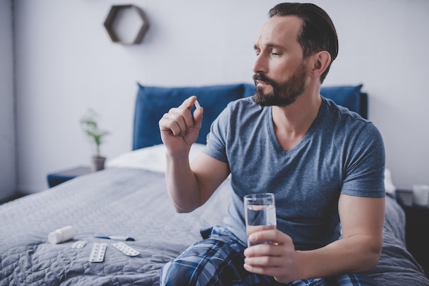 Caucasian man holding pill and glass of water sitting on the bed