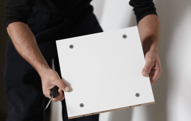 Caucasian man holding a white shelf assembling a cabinet close up of hands with a screwdriver Cropped image with selective focus Male in black clothing holding a white square board with four screws