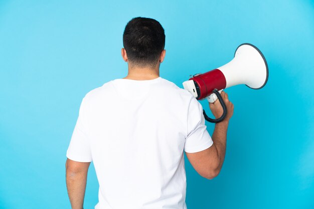 Caucasian man over isolated blue background holding a megaphone and in back position