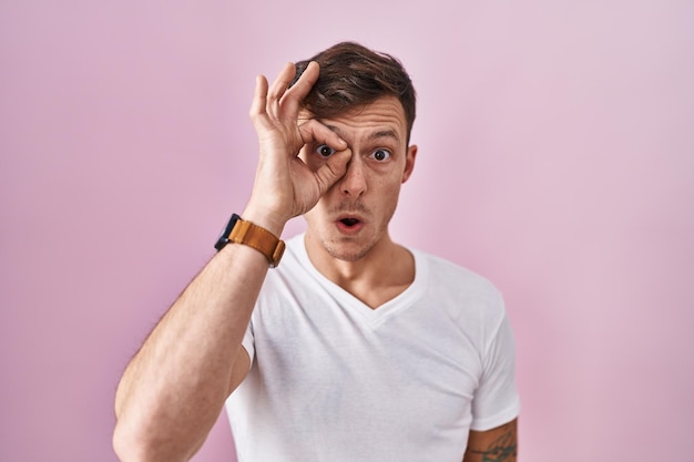 Caucasian man standing over pink background doing ok gesture shocked with surprised face eye looking through fingers unbelieving expression