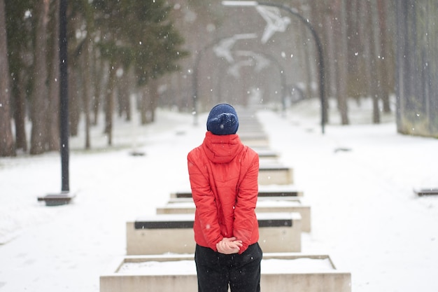 Caucasian man wearing red jacket enjoying snowfall in park, standing backfront