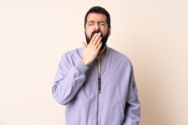 Caucasian man with beard wearing a jacket over isolated wall yawning and covering wide open mouth with hand