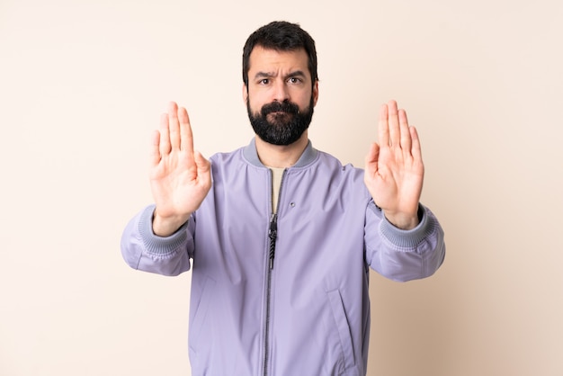 Caucasian man with beard wearing a jacket over wall making stop gesture and disappointed