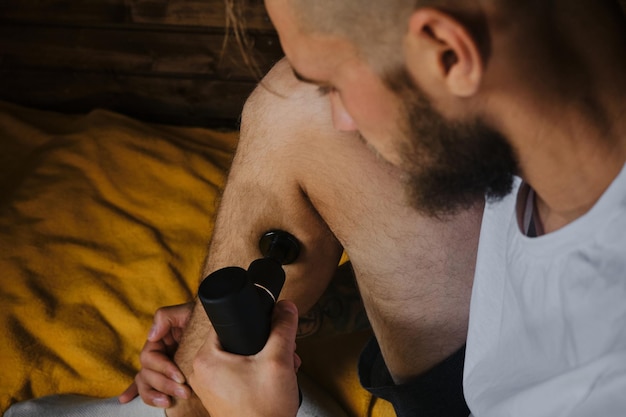 Caucasian man with dreadlocks gives himself massage with percussion gun at home sitting on bed