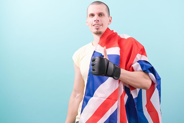 A caucasian MMA fighter in black gloves holds the UK flag and shows a thumb up on a blue background Occupation Discipline Courage Champion Up Male Fit Boxer Martial Ring Force Kick Posing