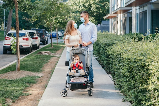 Photo caucasian mother and father in face masks walking with baby in stroller family strolling together