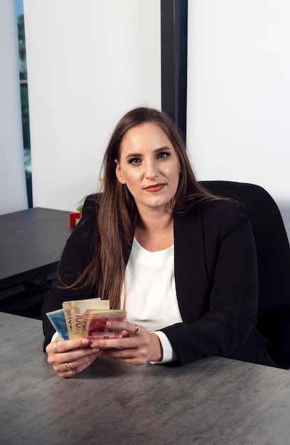 Caucasian office businesswoman in black suit holding money cash new shekels in hands Portrait of woman with long brunette hair holding fan of cash money in shekels banknotes and looking at camera