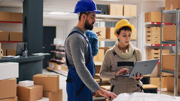 Caucasian people analyzing list of goods on tablet, working in warehouse with retail merchandise and boxes. Supervisor giving tasks to worker, organizing products on shelves and racks.