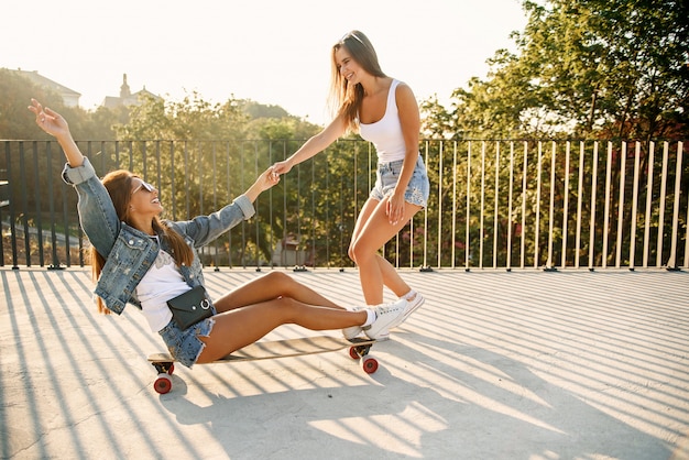 Caucasian pretty girl sits on a longboard while her girlfriend goes forward and holds her hand.