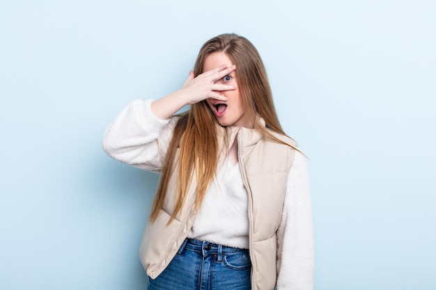 Caucasian red hair woman looking shocked, scared or terrified, covering face with hand and peeking between fingers