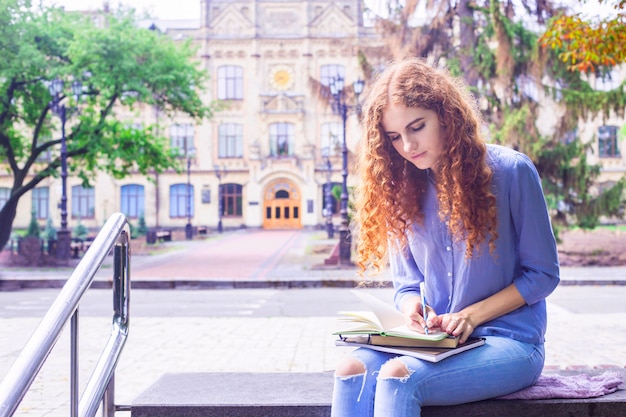 A Caucasian red-haired, curly-haired girl sits near her university 