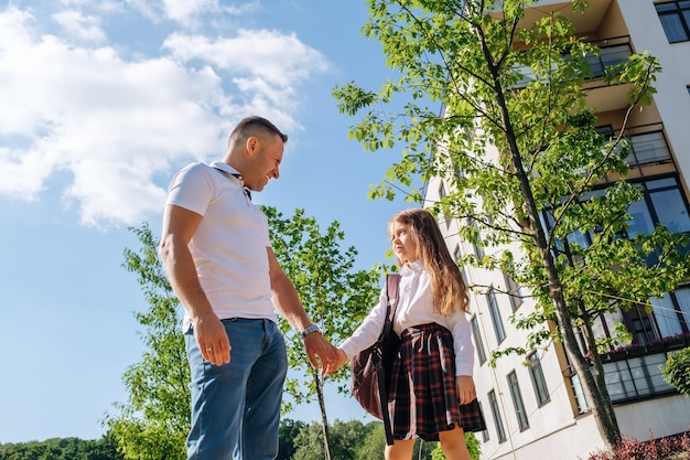 Caucasian schoolgirl with a backpack and uniform holds her smiling father hand