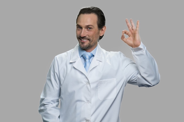 Caucasian scientist man showing ok with fingers. Portrait of cheerful middle-aged man in lab coat looking at camera on gray background.