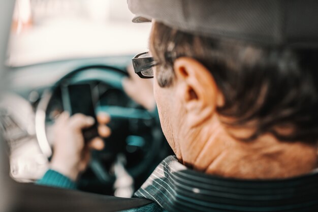 Caucasian senior man with cap on head and eyeglasses sitting in car with hand on steering wheel and using smart phone.