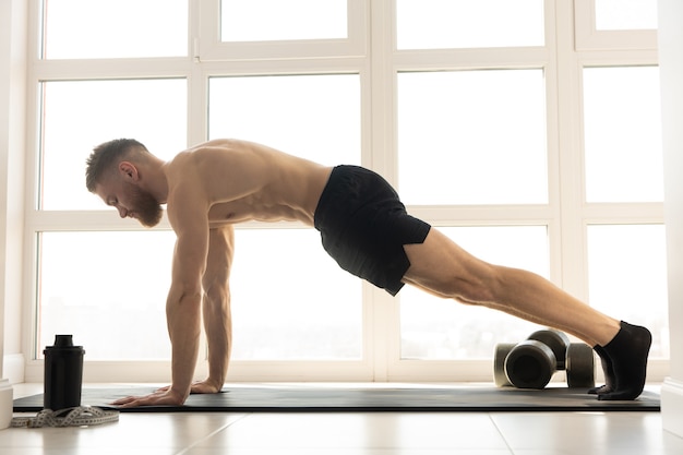 Caucasian sportsman doing plank for abdominal muscles on fitness mat. Young bearded man with naked torso. Concept of sport activity at home. Interior of modern spacious apartment. Sunny daytime