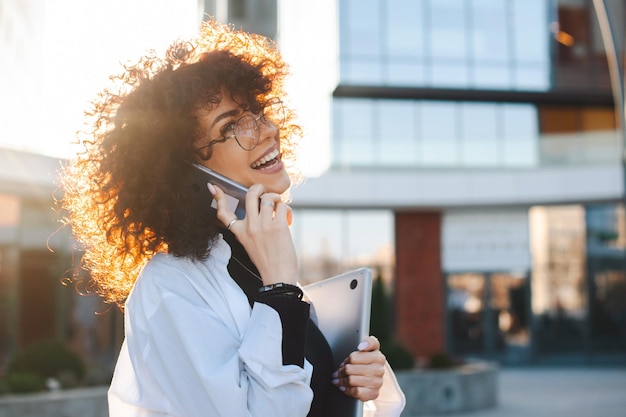 Caucasian student woman talking on the phone and smiling while walking outdoor curly haired business