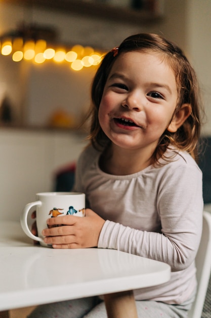 Caucasian toddler girl drinking cocoa from mug at home. Christmas bokeh lights. High quality photo