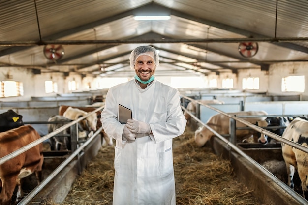 Caucasian unshaven veterinarian in protective uniform standing in barn with tablet under armpit.